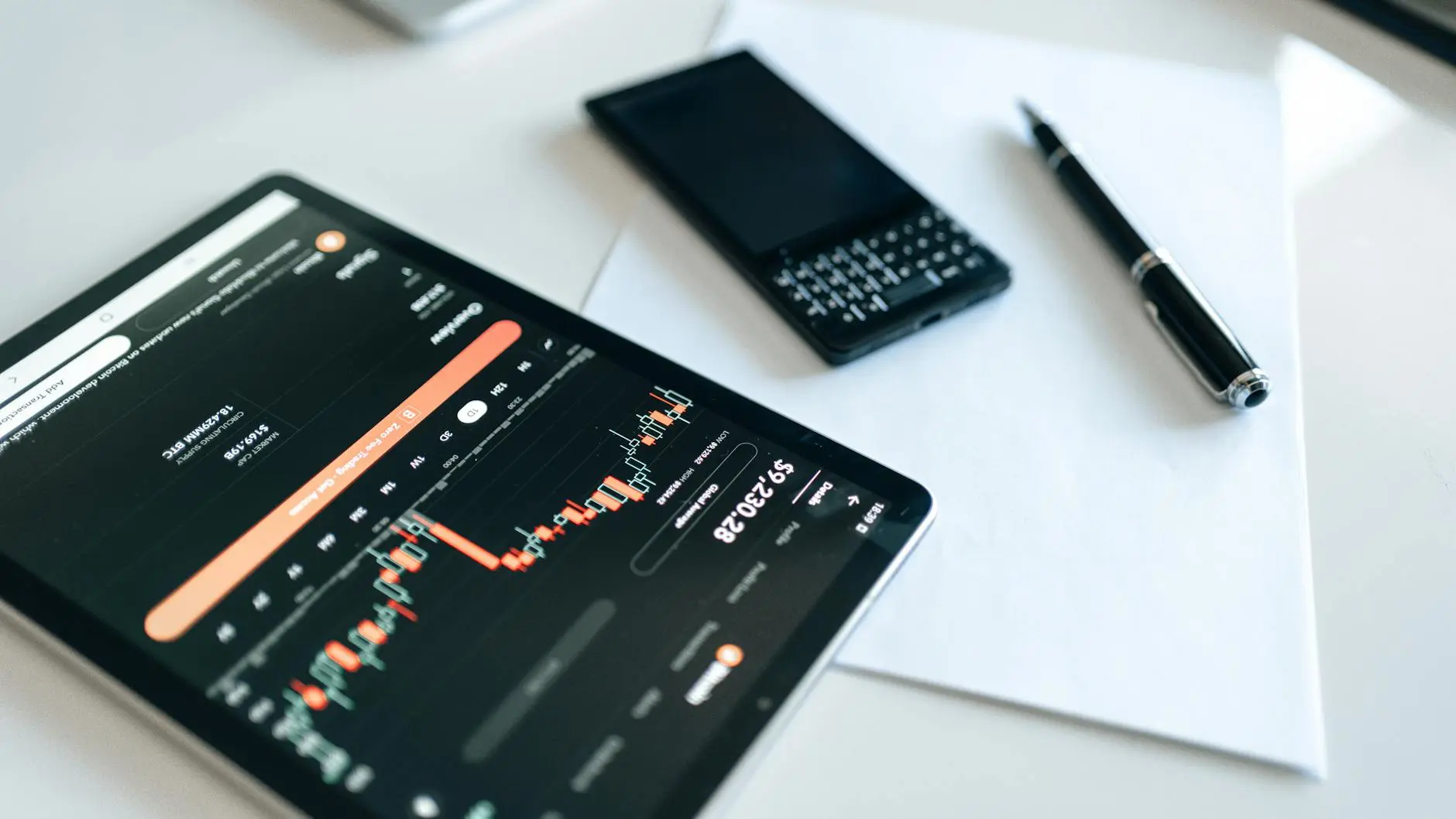 A cryptocurrency trading chart displayed on a tablet with a smartphone and pen.