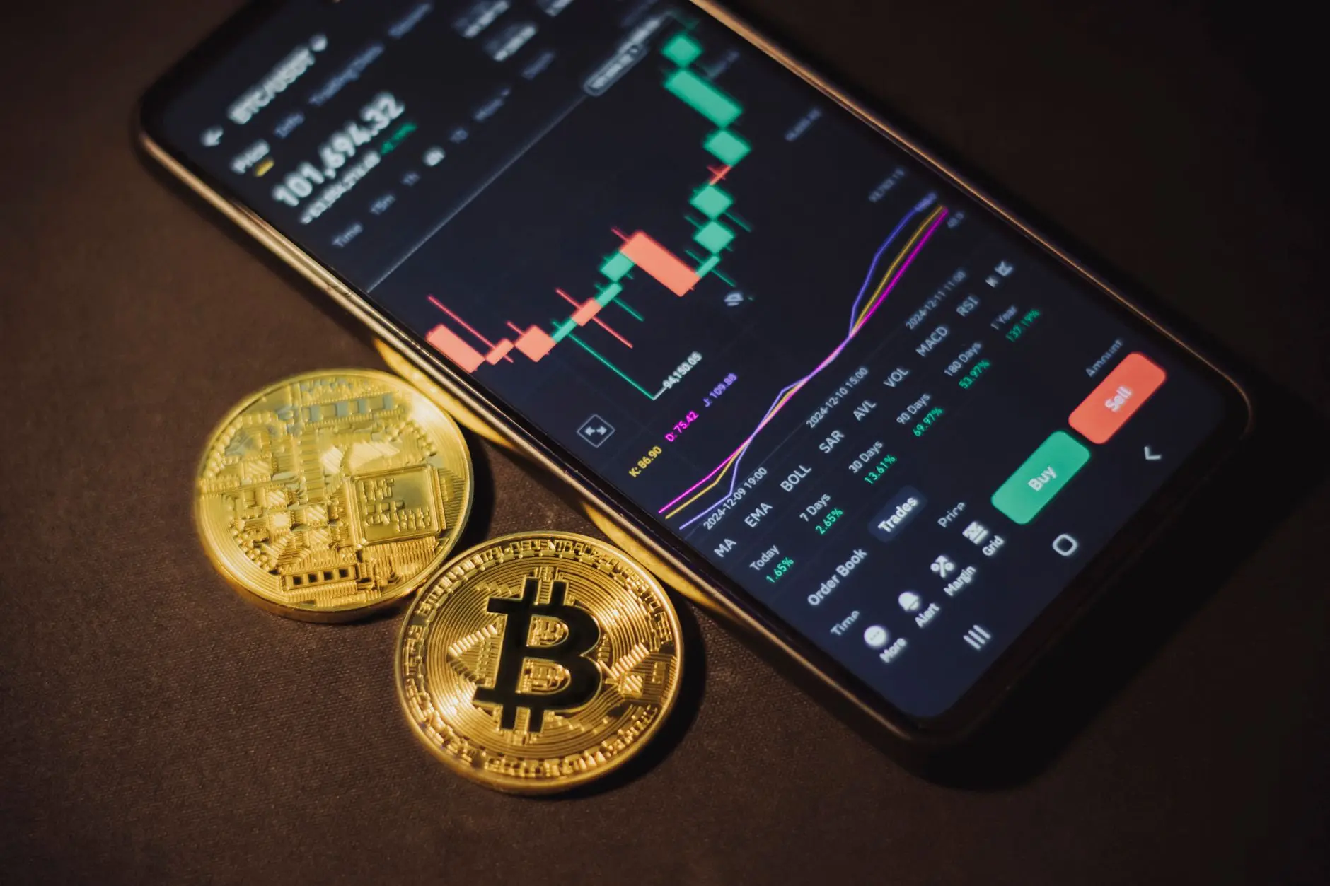 A smartphone displaying a bitcoin trading app alongside Bitcoin coins.