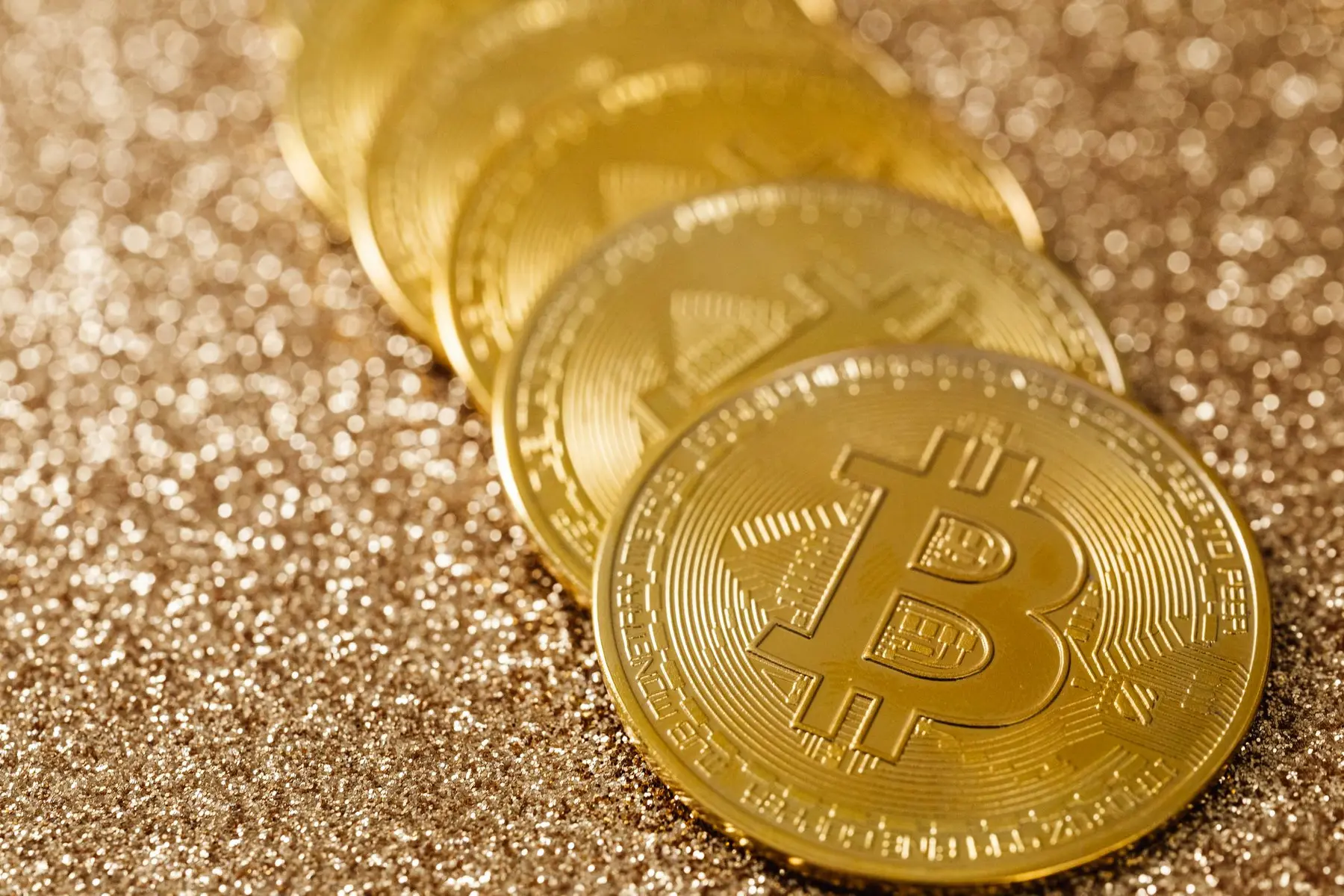 A row of gold Bitcoin physical tokens resting on a shimmering gold glitter surface