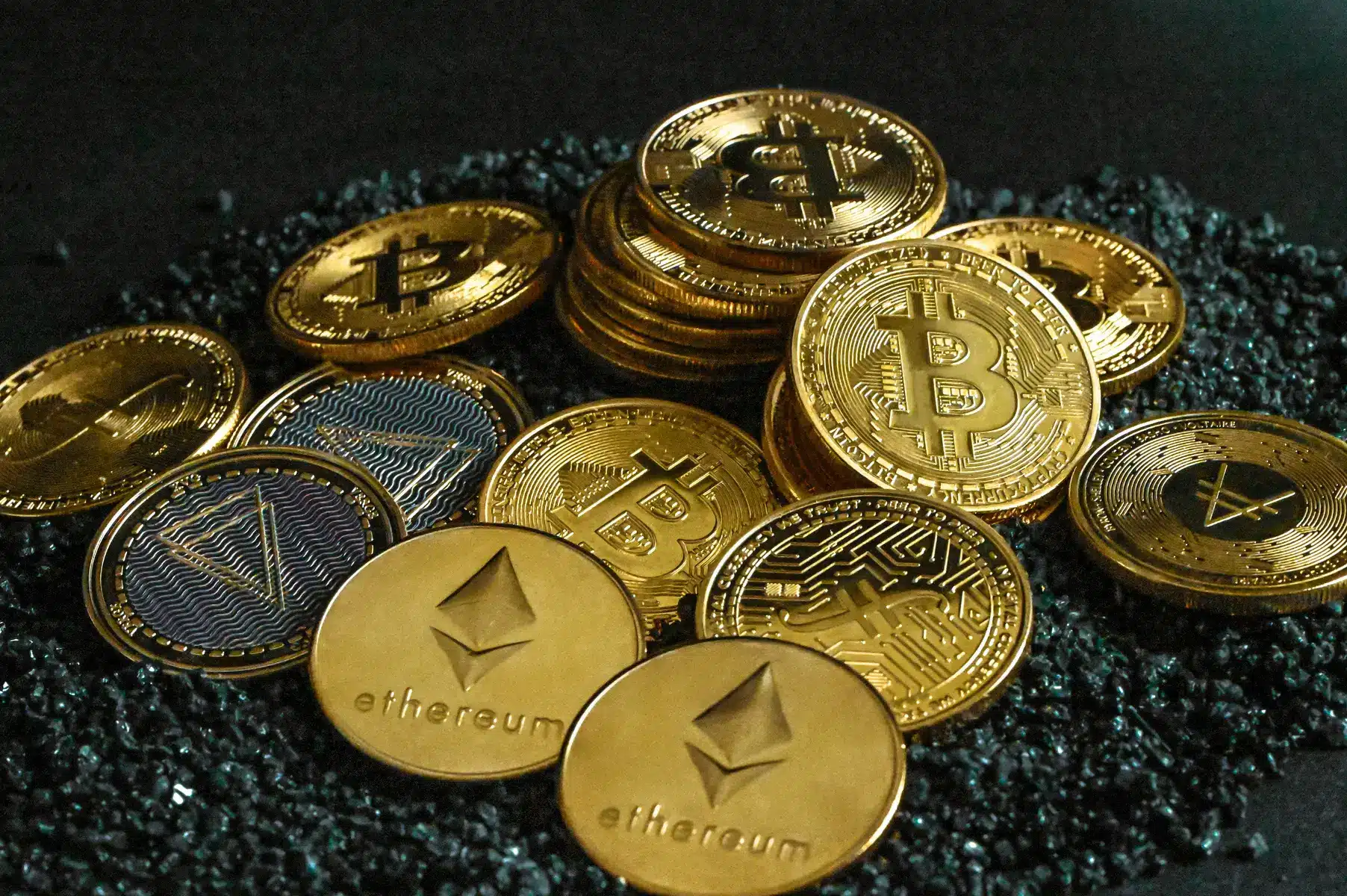 Goldene Kryptowährungs-Münzen, darunter Bitcoin und Ethereum, auf einem dunklen, steinigen Untergrund.