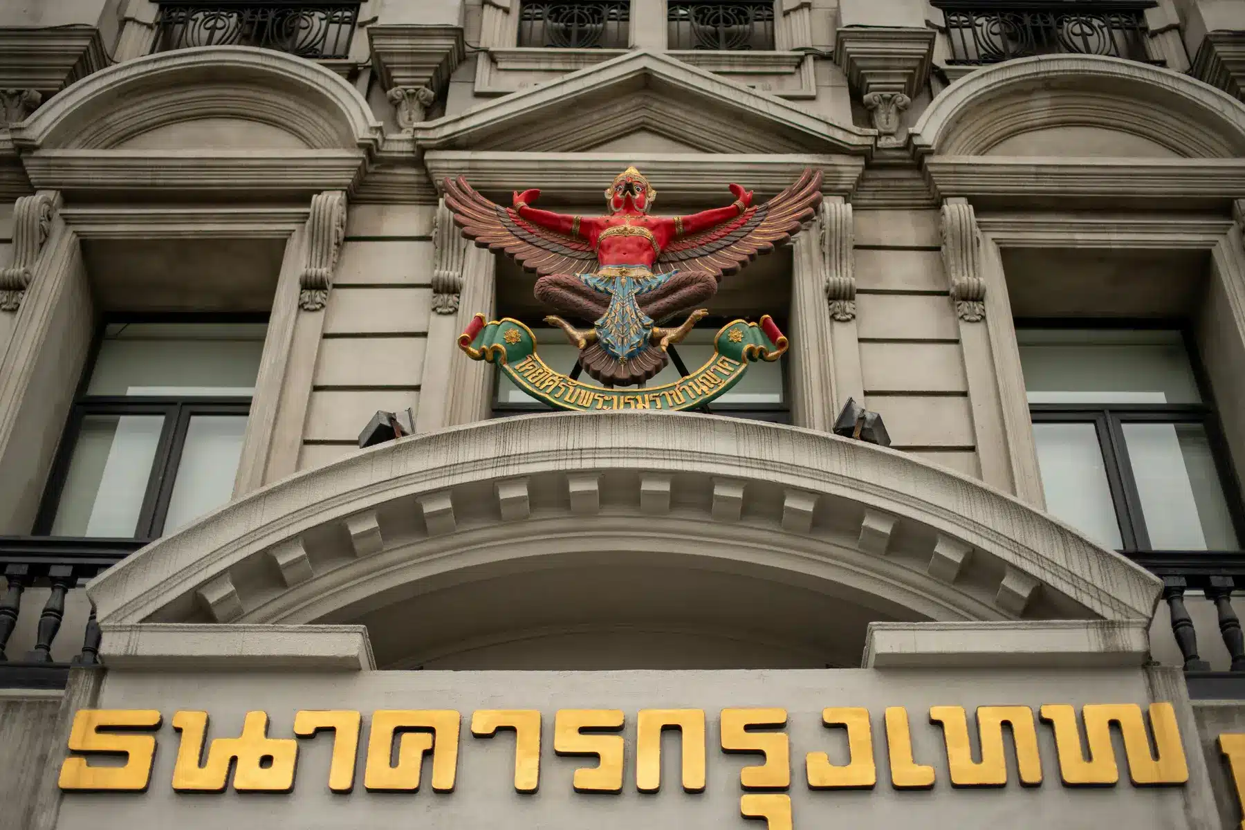 Detail der Steinfassade des Bangkok Bank Gebäudes mit einem goldenen Emblem und thailändischer Schrift.
