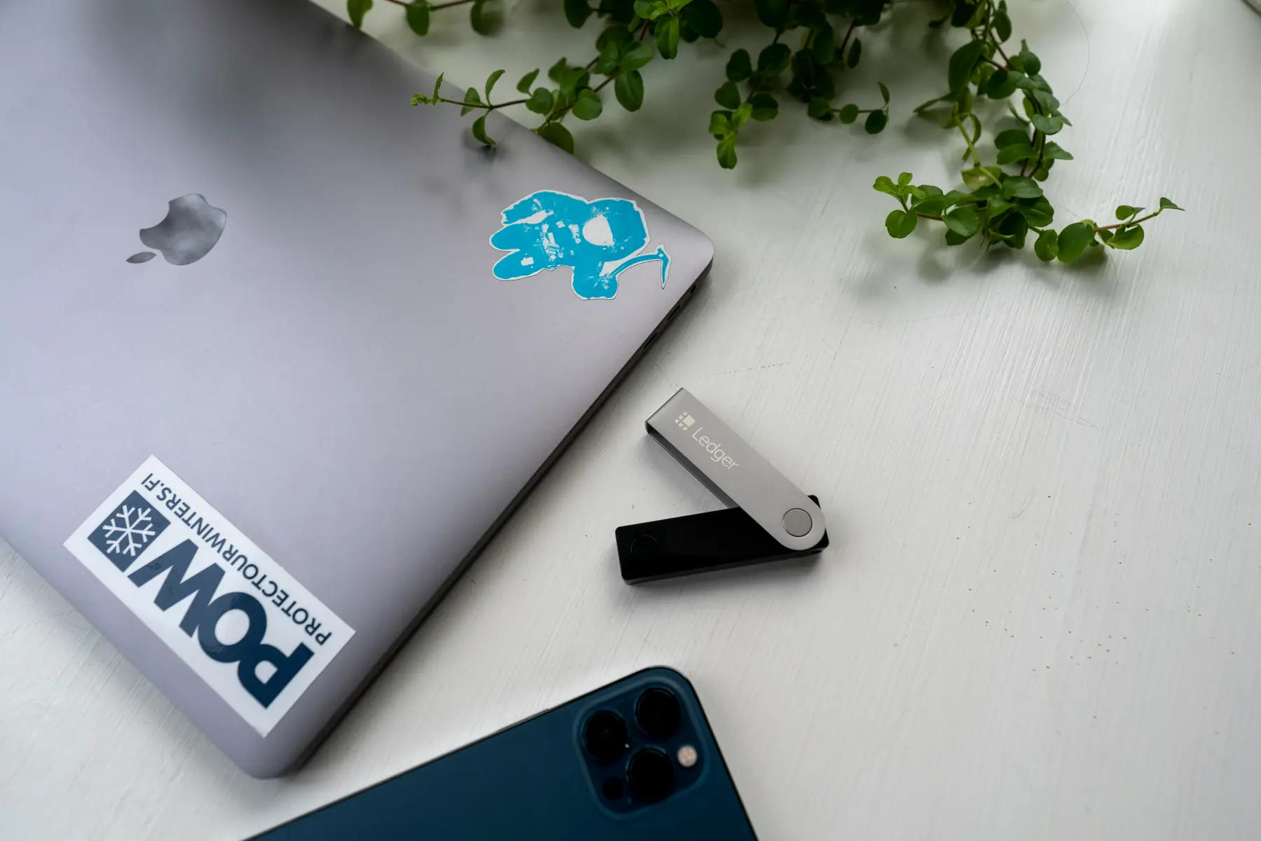 Ein Ledger Hardware-Wallet neben einem MacBook und einem iPhone auf einem weißen Tisch.