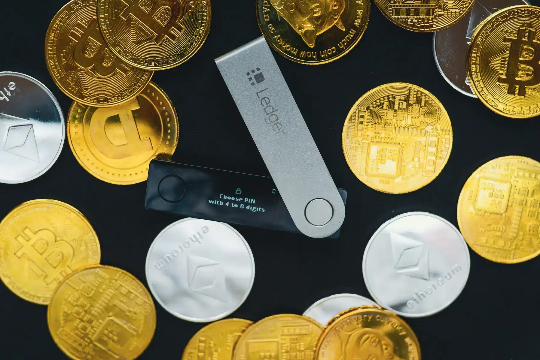 Ein Ledger Hardware-Wallet neben verschiedenen physischen Kryptowährungs-Münzen auf schwarzem Hintergrund.