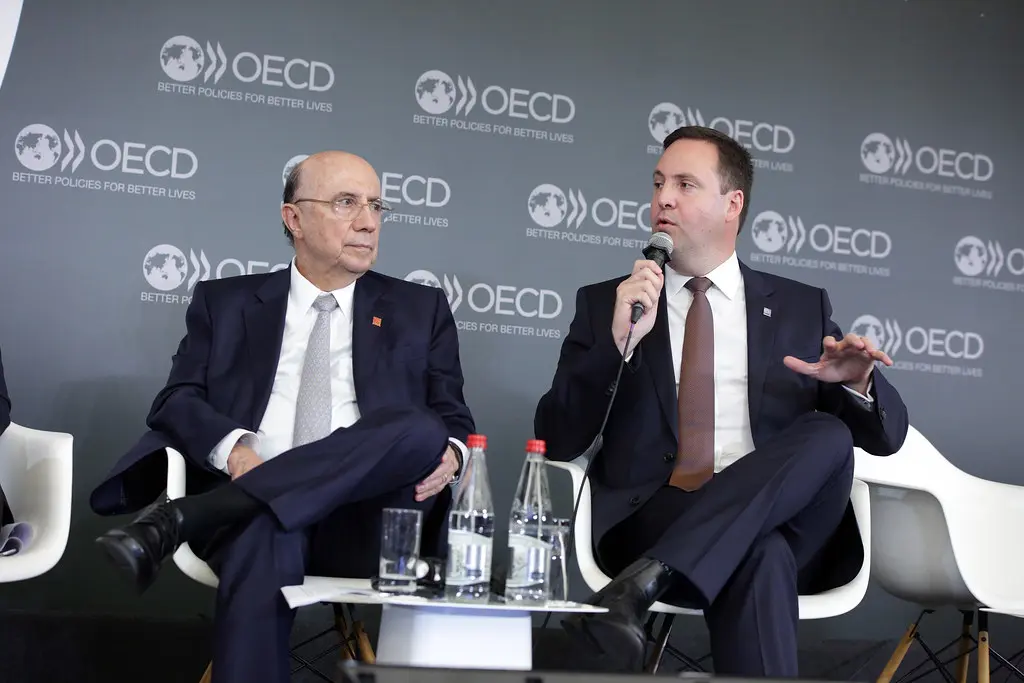 Twee mannen in pakken tijdens een gesprek tegen een achtergrond met het logo van de OECD.
