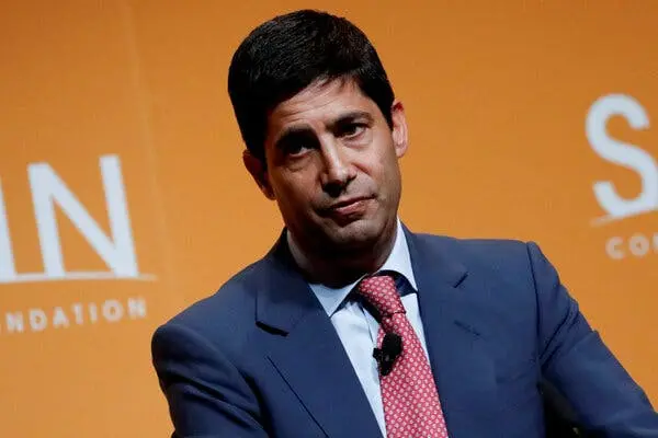 Portrait of Kevin Warsh wearing a blue suit and red patterned tie against an orange background