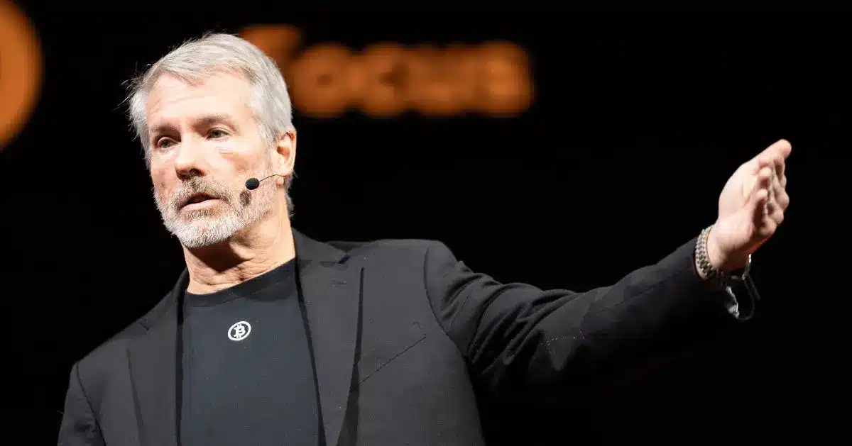 Michael Saylor speaking at a podium wearing a black blazer and a Bitcoin logo t-shirt