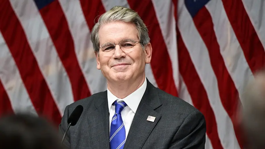 Portrait of Scott Bessent in a dark suit and blue tie standing before American flags.